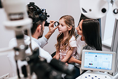 Optometrist examining a young girl’s eyes while she sits on her mother’s lap 